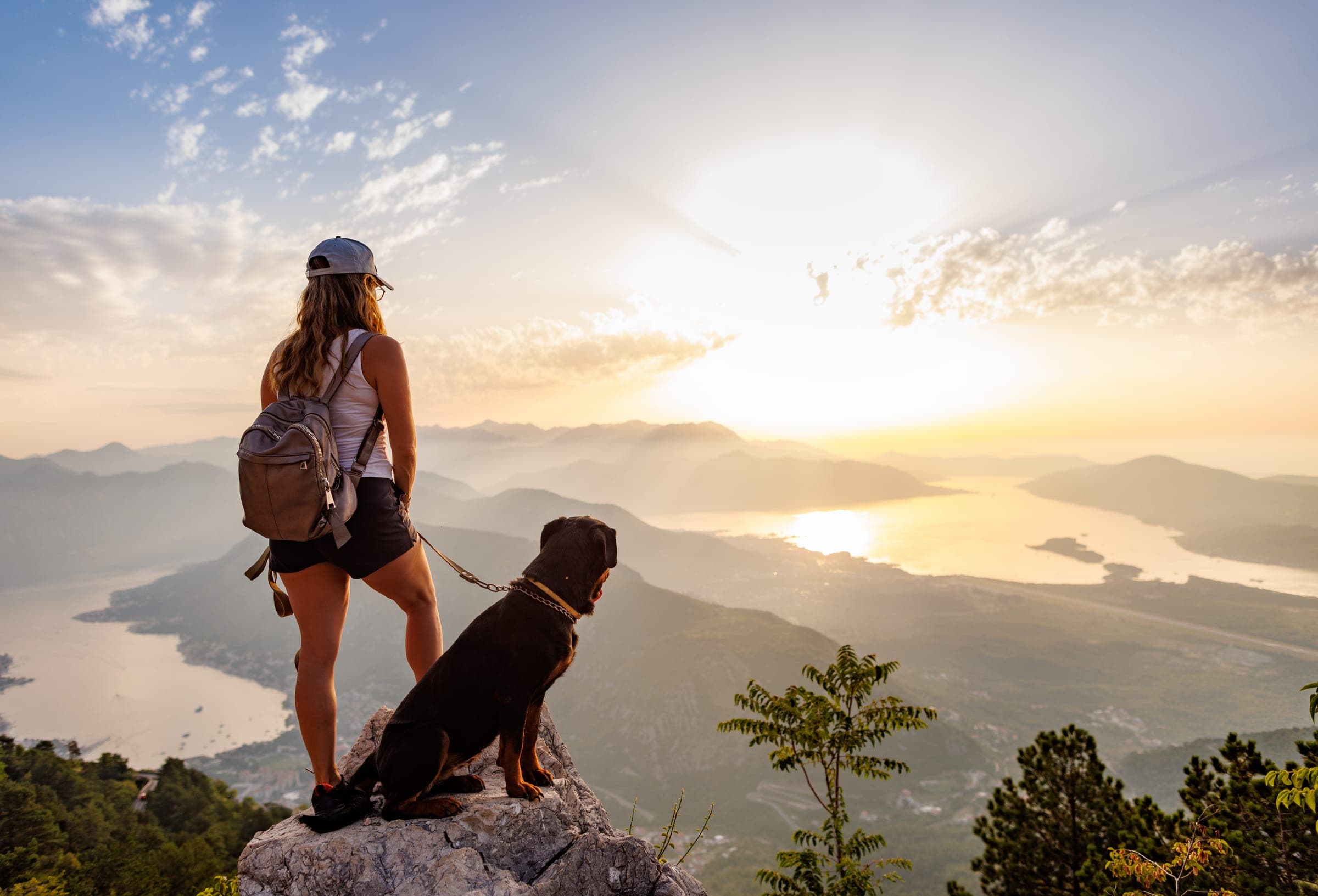 A sporty hiker with a backpack standing at the edge of a mountain overlook beside a Rottweiler.